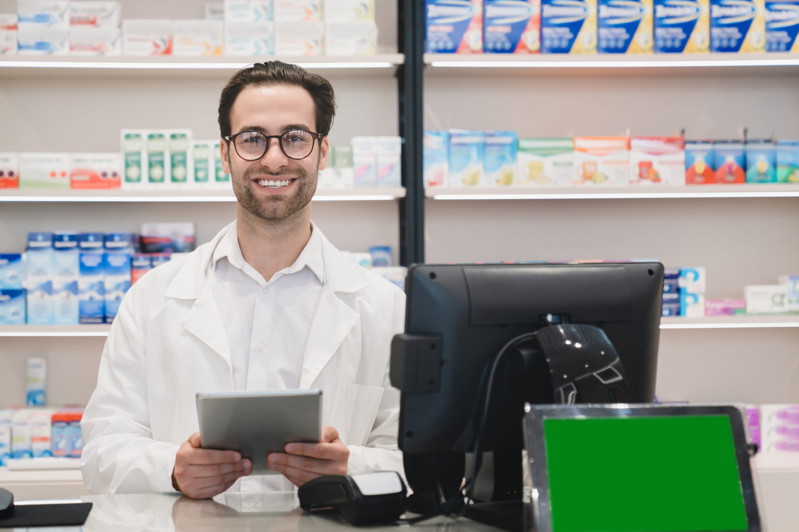 Confident smiling male caucasian druggist pharmacist using digital tablet standing at cash point desk in pharmacy drugstore with green screen with mockup copyspace
